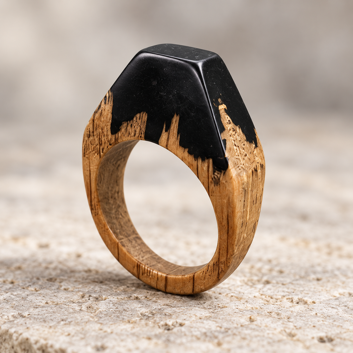A close-up of a ring with a natural wood band and a black top section featuring a textured, brushstroke-like pattern, set against a neutral, sandy background. Leseni prstan, leseni nakit,