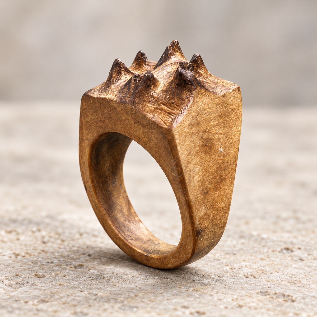 A rustic, textured metallic ring with a crown-like spiked top, displayed on a neutral stone surface.