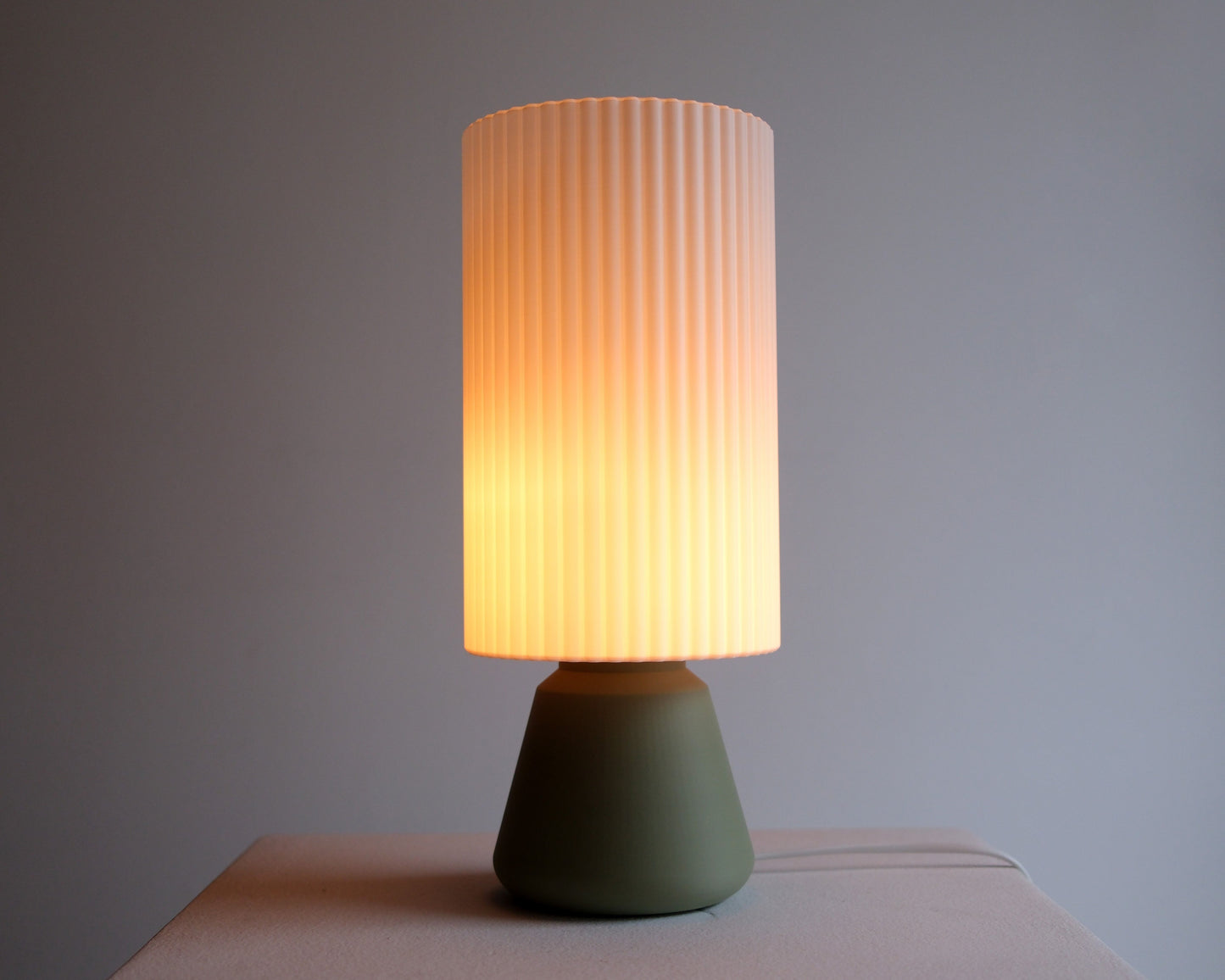 A modern table lamp with a ribbed pleated shade that glows with a warm yellow light, set on a matte olive green base against a neutral gray background.