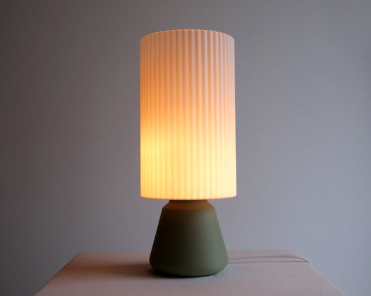 A modern table lamp with a ribbed pleated shade that glows with a warm yellow light, set on a matte olive green base against a neutral gray background.