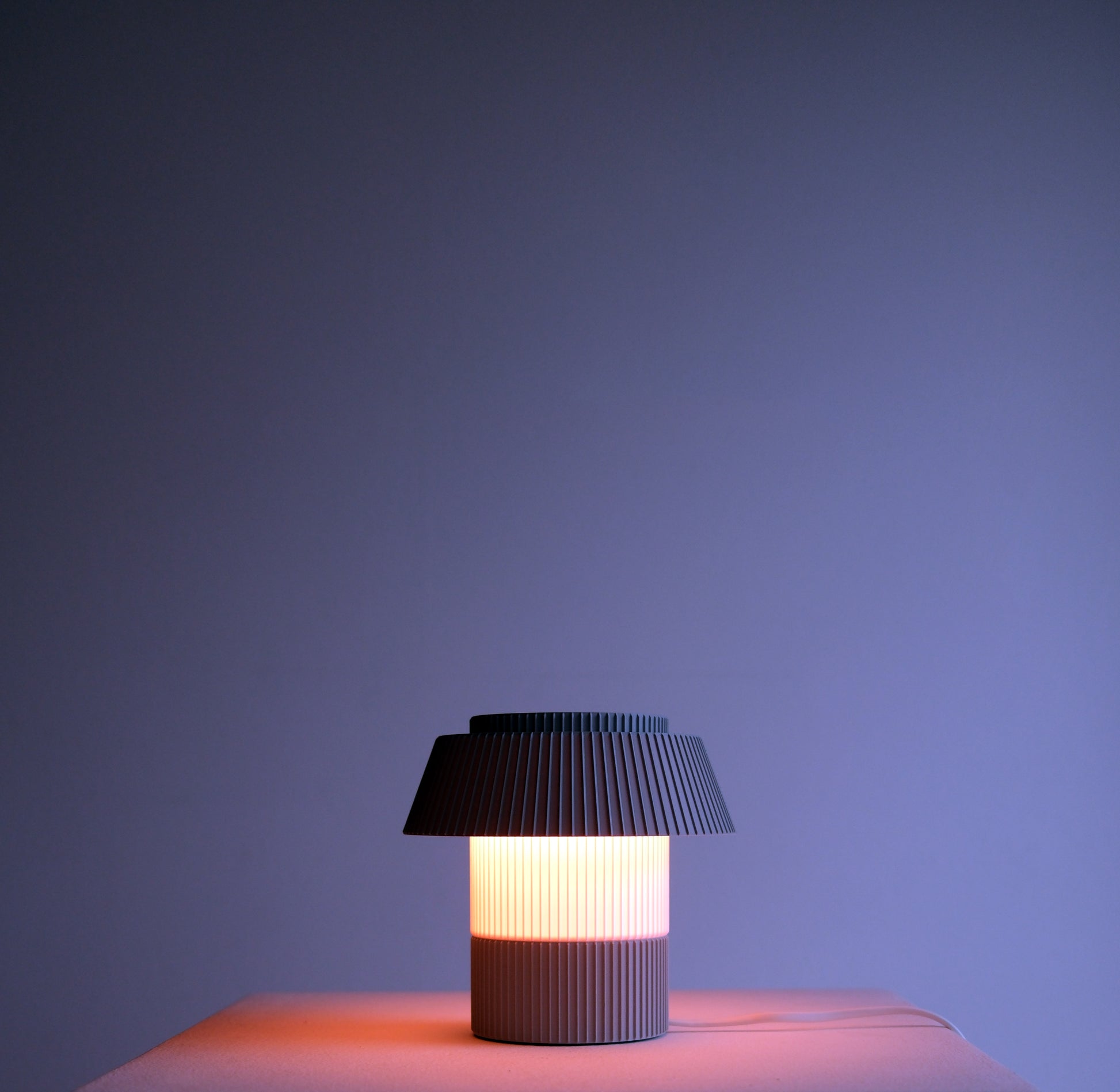 A modern pleated table lamp with a dark gray pleated shade and a ribbed cylindrical base, glowing with a warm light against a dark blue-purple background.