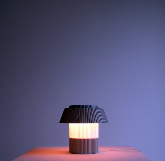 A modern pleated table lamp with a dark gray pleated shade and a ribbed cylindrical base, glowing with a warm light against a dark blue-purple background.