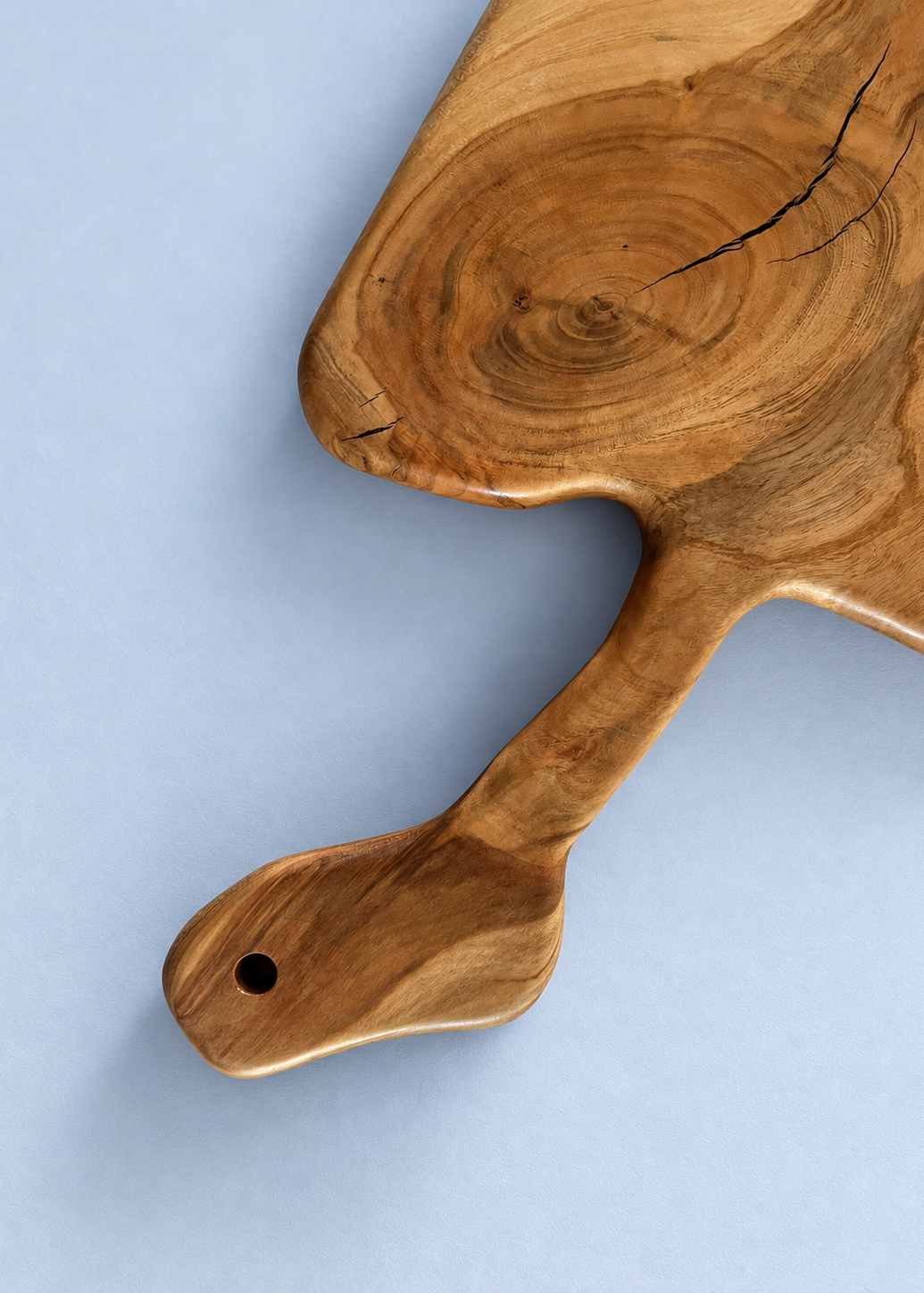 A close-up of a handcrafted wooden serving board with a natural live edge and handle, shown on a light blue background.