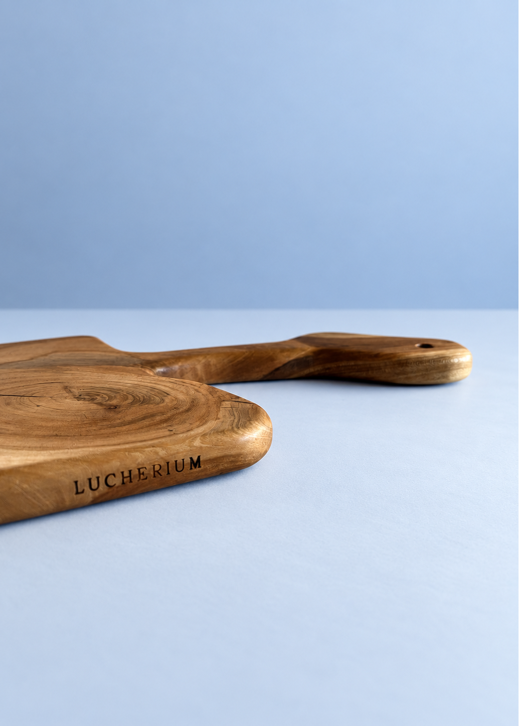 A close-up of a wooden serving board with a handle, featuring the engraved brand name 'LUCHERIUM' on a light blue background.
