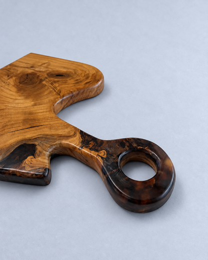 A rustic wooden serving board with a decorative handle, featuring a light honey-toned wood body and a darker, highly figured tortoiseshell-like end, photographed on a neutral light gray background.