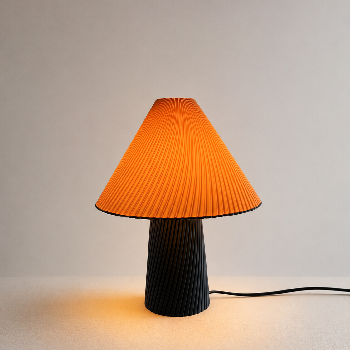 A pleated table lamp with a bright orange conical shade and a ribbed black base, shown lit against a plain light gray background.