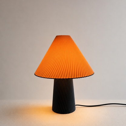 A pleated table lamp with a bright orange conical shade and a ribbed black base, shown lit against a plain light gray background.