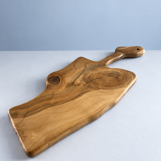 A rustic wooden serving board with a natural live edge, grain pattern, and a small handle with a hanging hole, photographed on a light gray surface against a blue-gray background.