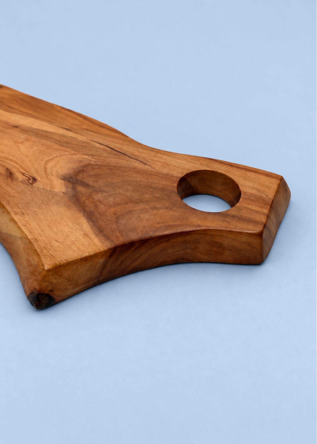 A close-up of a wooden serving board or cutting board handle with a natural live edge and a circular hole, shown on a light blue background.